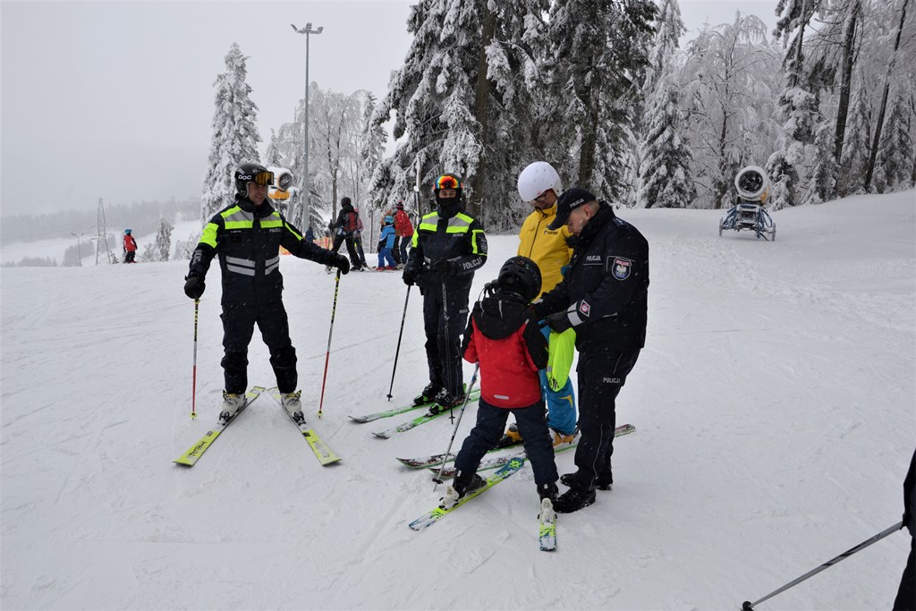 na szczycie stoku policjanci rozmawiają z chłopcem na temat bezpiecznej jazdy na nartach, jeden z nich wręcza mu element odblaskowy