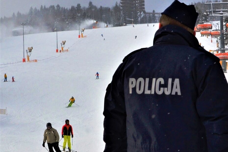 na pierwszym planie policjant prewencji odwrócony tyłem, w tle narciarze zjeżdżający ze stoku