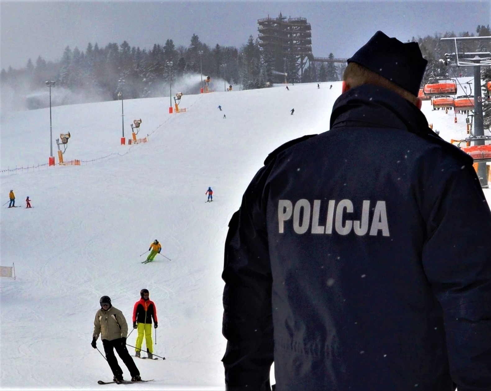 na pierwszym planie policjant prewencji odwrócony tyłem, w tle narciarze zjeżdżający ze stoku